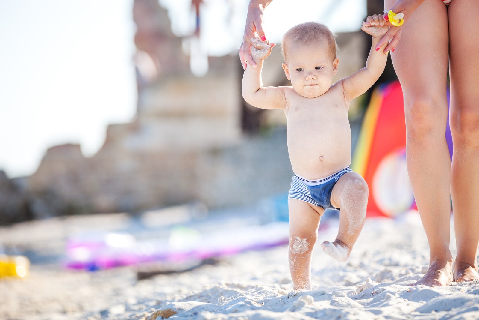 Baby boy taking first steps in the sand at Bondi Beach, Australia, supported by his mother, with a bright beach backdrop and clear summer light.