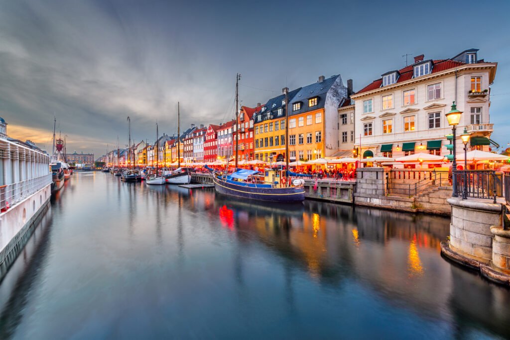 Vibrant evening view of Copenhagen’s Nyhavn canal with colorful historic buildings, docked boats, and lively waterfront cafes glowing under twilight.