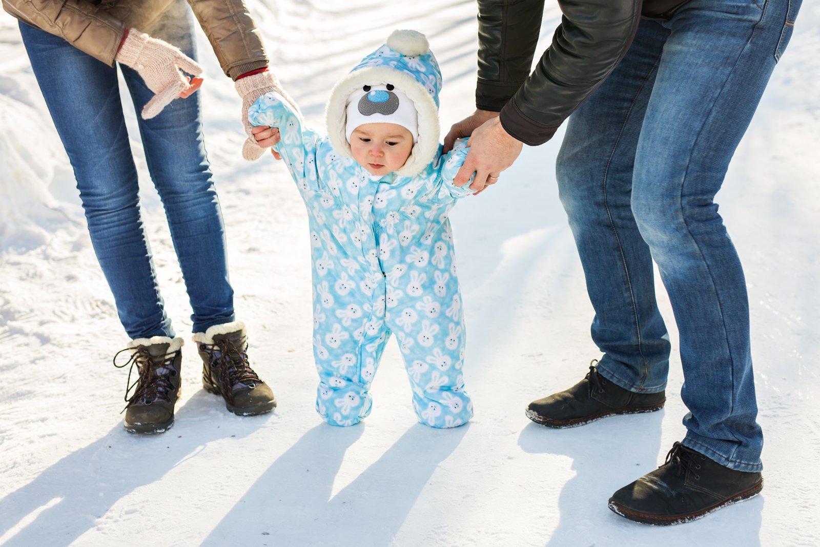 Baby bundled in a blue bunny-print snowsuit taking first steps on a snowy path in Reykjavik, Iceland, supported by parents in winter boots.