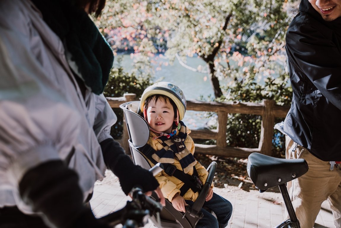 Happy toddler wearing a helmet sitting in a child bike seat during a family biking trip in a park in Tokyo, Japan, with autumn trees and a scenic riverside in the background.