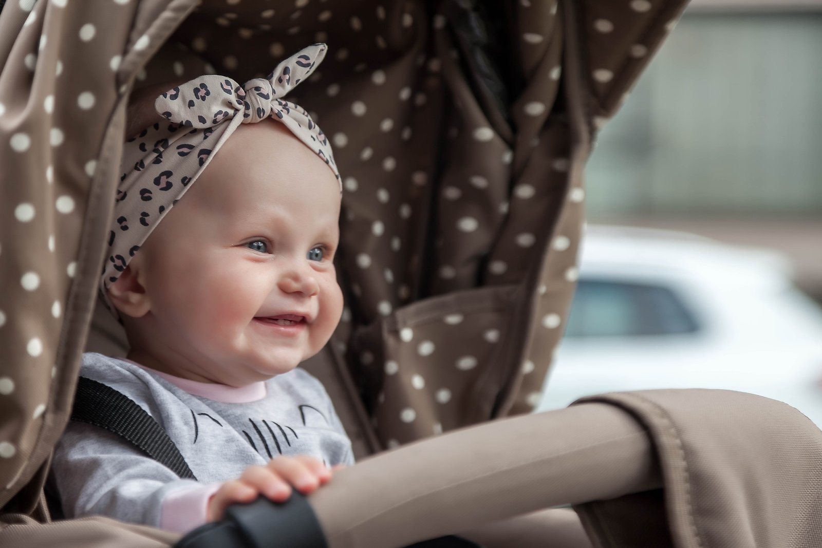 Smiling baby girl in a stroller with a polka-dot canopy and leopard-print headband, enjoying a stroll through the stroller-friendly streets of Amsterdam, Netherlands.